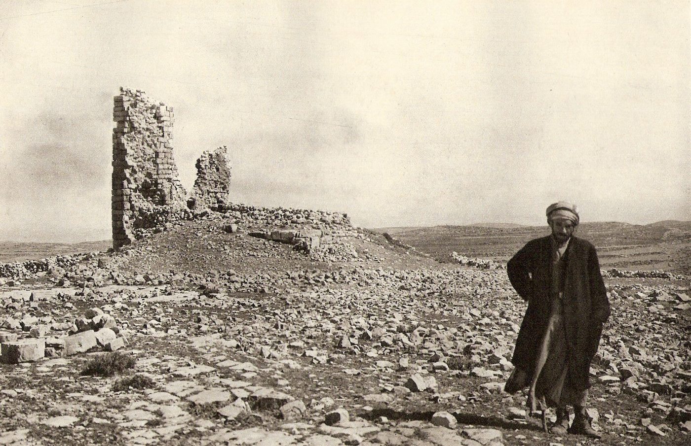 Black-and-white 1925 view of the ruins of Beth Zur, with a standing wall on a rocky hill and a figure in the foreground.